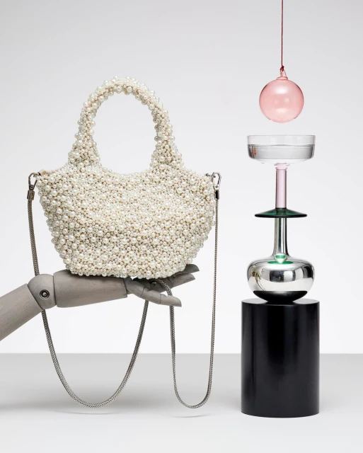 Pearl-encrusted handbag held by mannequin hand next to artistic glassware on a cylindrical pedestal in a minimalist studio setting.
