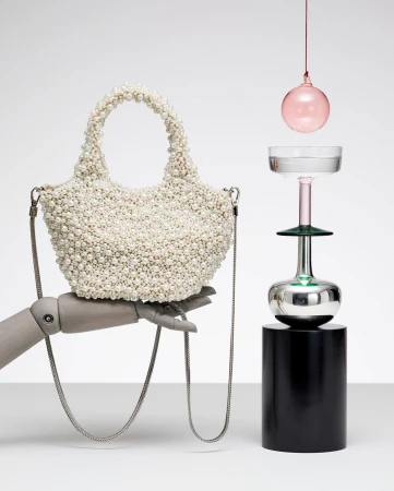 Pearl-encrusted handbag held by mannequin hand next to artistic glassware on a cylindrical pedestal in a minimalist studio setting.