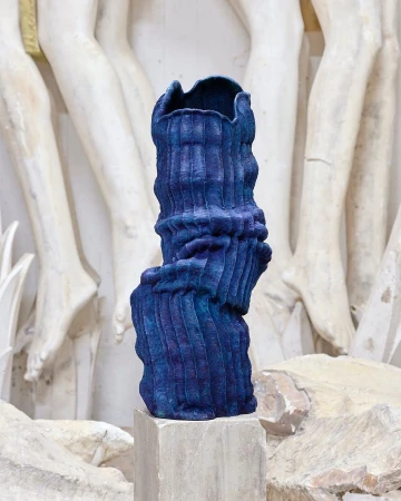 Striking blue textured vase on a pedestal against a sculpted wall in a studio setting. Modern art with classical elements.