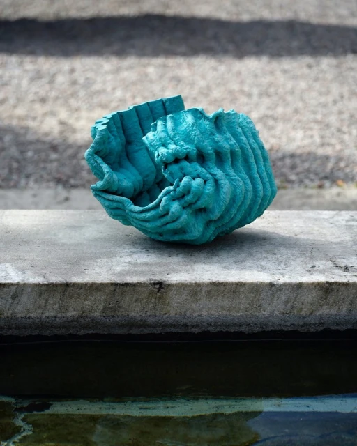 Sculptural turquoise art piece with layered textures on a concrete ledge with a water feature in the background, outdoor setting.