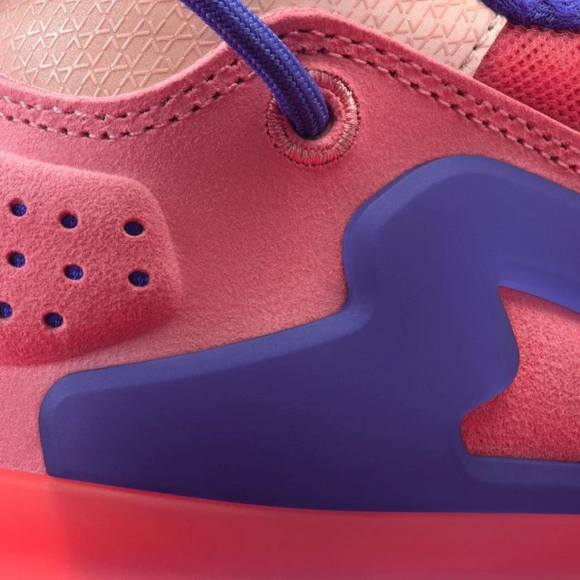 Close-up shot of a stylish sneaker featuring pink suede, a purple rubber accent, and blue laces, highlighting the shoe's design and texture.