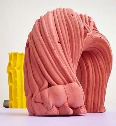 Abstract pink sculpture with linear texture and a yellow geometric form behind it, set against a neutral white background.