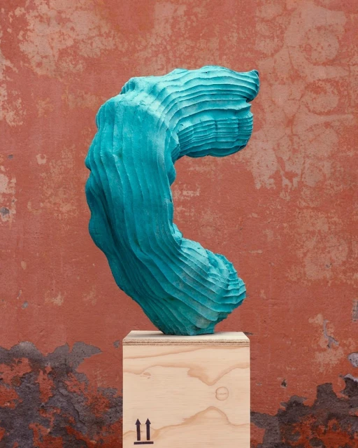 Turquoise sculpture on a wooden crate in front of a textured orange wall, showcasing contemporary art and minimalist design.