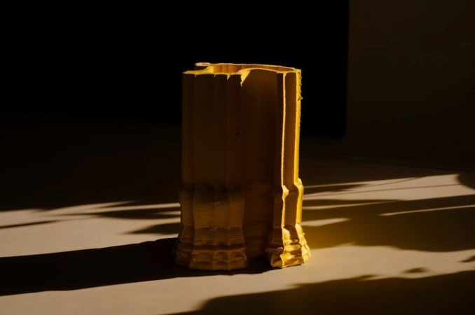 Abstract yellow sculpture on a neutral surface with shadows, featuring geometric shapes and a textured finish under studio lighting.