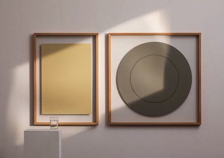 Two framed abstract artworks hang on a wall next to a small white pedestal table with a glass of water in a well-lit room.