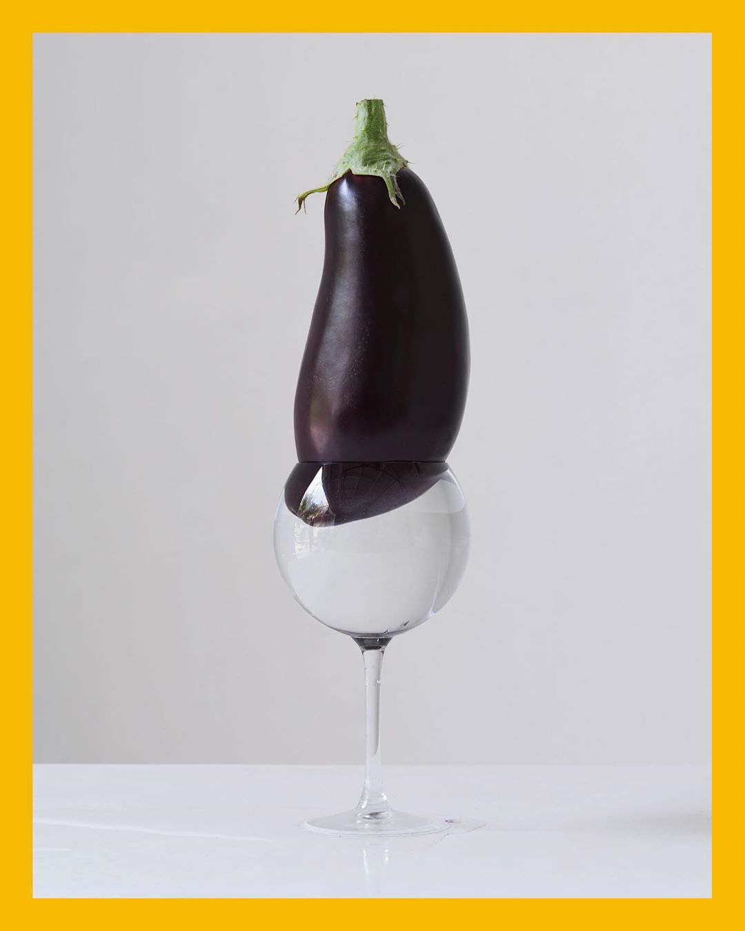 Still life of an eggplant balanced on top of a clear wine glass filled with water, against a neutral background with a yellow border.