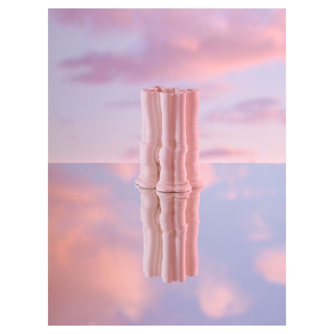 A light pink vase reflects in a glossy surface, set against a soft pink and blue sky background with gentle cloud formations.