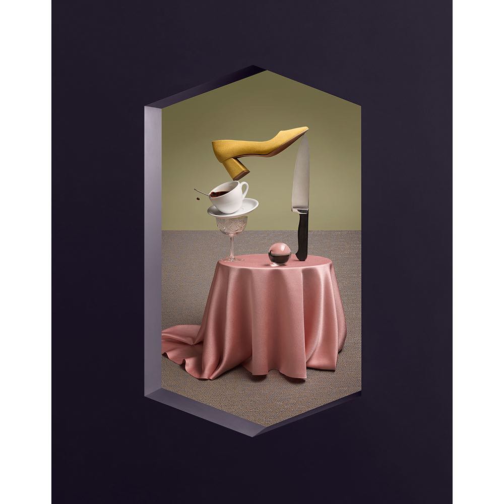 Still life composition features a yellow pump shoe, coffee cup, knife, and ball on a table with a pink tablecloth against a green background.