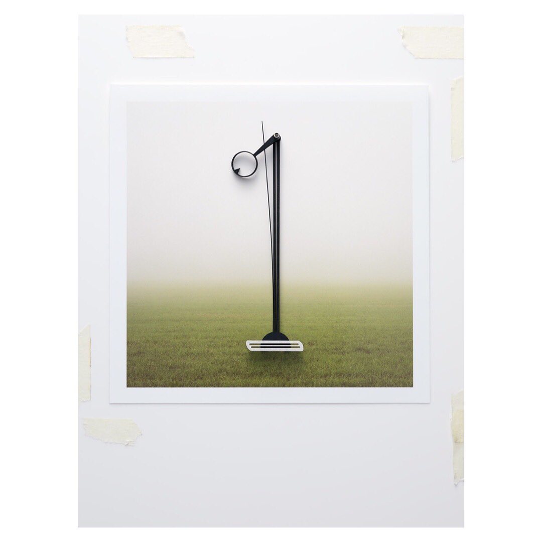 Contemporary photo of a minimalist black floor lamp positioned on a white framed image with a grass field and fog background, indoor setting.