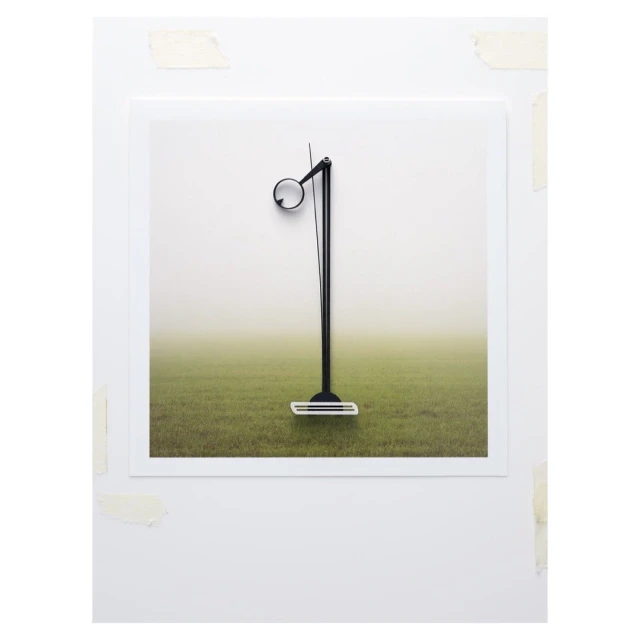 Contemporary photo of a minimalist black floor lamp positioned on a white framed image with a grass field and fog background, indoor setting.