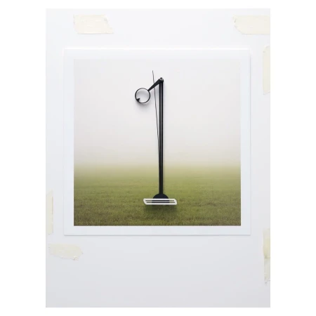 Contemporary photo of a minimalist black floor lamp positioned on a white framed image with a grass field and fog background, indoor setting.