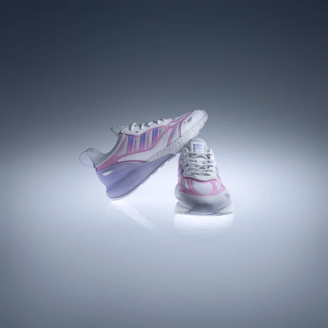 Two Adidas ZX 2K Boost 2.0 sneakers, one balancing on the other in a studio setting with a gradient gray and white background.