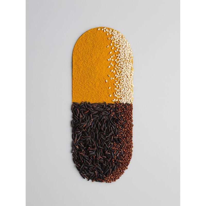 Pill-shaped mound of turmeric, sesame seeds, wild rice, and brown quinoa on a gray surface, arranged to mimic a capsule.