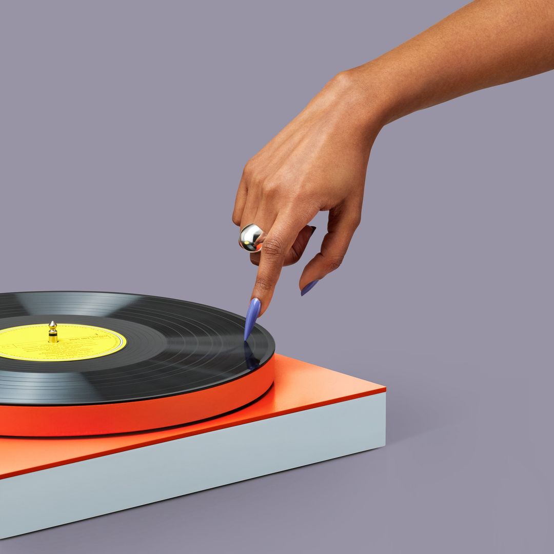 Vinyl record on a turntable, being selected by a woman's hand with a silver ring and lilac nails, against a grey background.