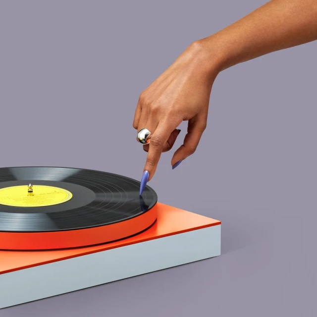 Vinyl record on a turntable, being selected by a woman's hand with a silver ring and lilac nails, against a grey background.