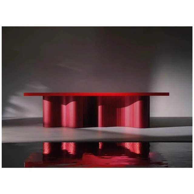 Eye-level shot of a modern red table with vertical slats in a minimalistic studio setting. Reflections are visible on the floor.