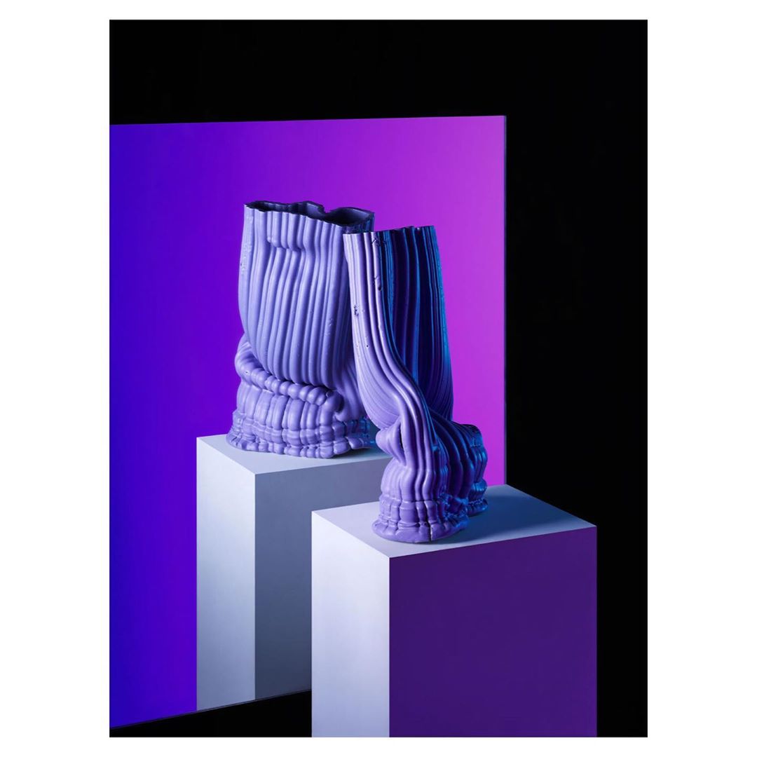Two purple textured vases on white pedestals against a split purple and black background in a studio setting with blue lighting.