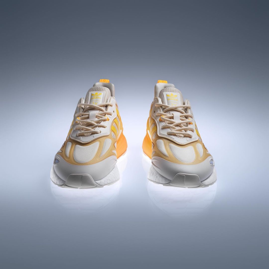 Two yellow and white Adidas ZX 2K Boost 2.0 sneakers are displayed on a light grey reflective surface, shot in a studio setting.
