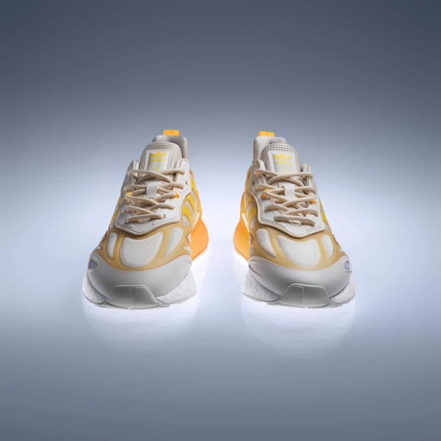 Two yellow and white Adidas ZX 2K Boost 2.0 sneakers are displayed on a light grey reflective surface, shot in a studio setting.