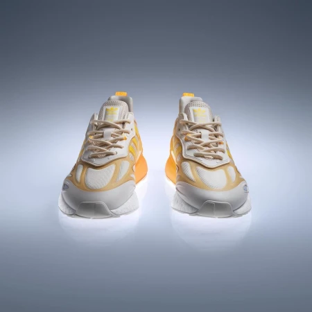 Two yellow and white Adidas ZX 2K Boost 2.0 sneakers are displayed on a light grey reflective surface, shot in a studio setting.
