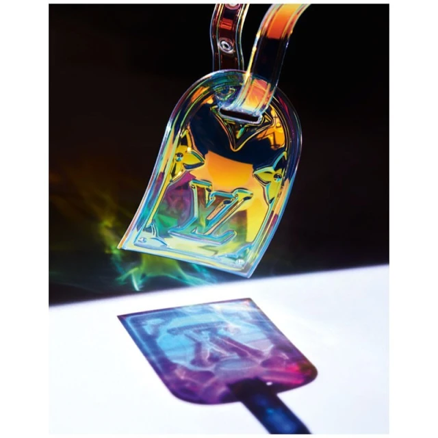 Close-up of a transparent LV-branded bag charm reflecting light, set against a gradient black to white background, product shot.