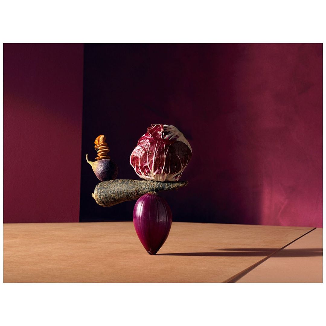 Still life featuring a balanced arrangement of purple onion, carrot, radicchio, and stacked figs against a plum and brown backdrop.