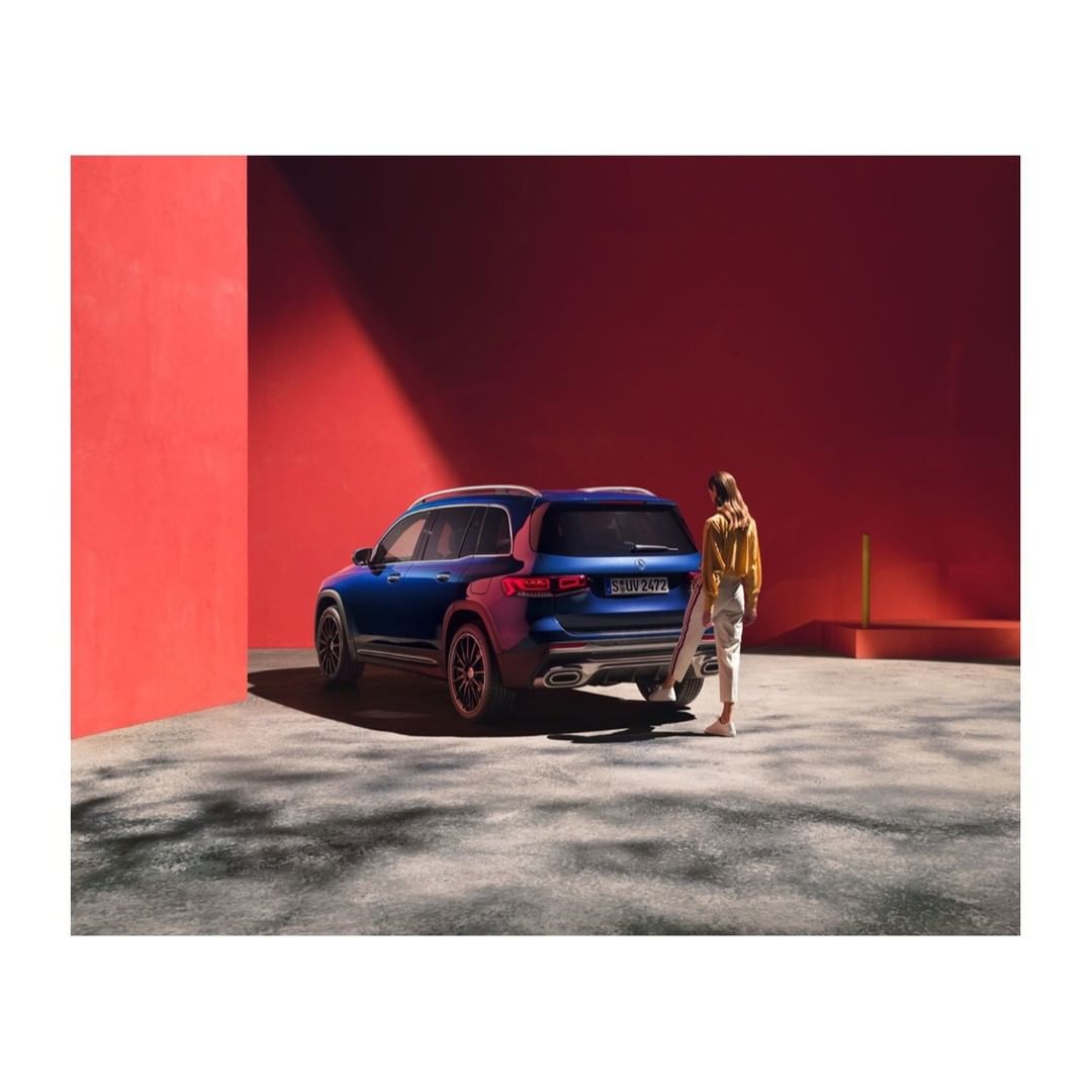Blue SUV parked against a bright red architectural wall with a woman standing beside it in a courtyard.