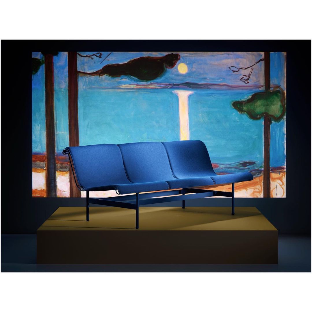 Blue fabric three-seater sofa with black metal frame on a raised yellow platform, set against a dark backdrop with a colorful painting.