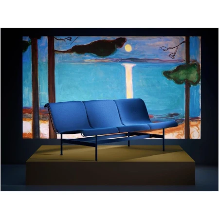 Blue fabric three-seater sofa with black metal frame on a raised yellow platform, set against a dark backdrop with a colorful painting.
