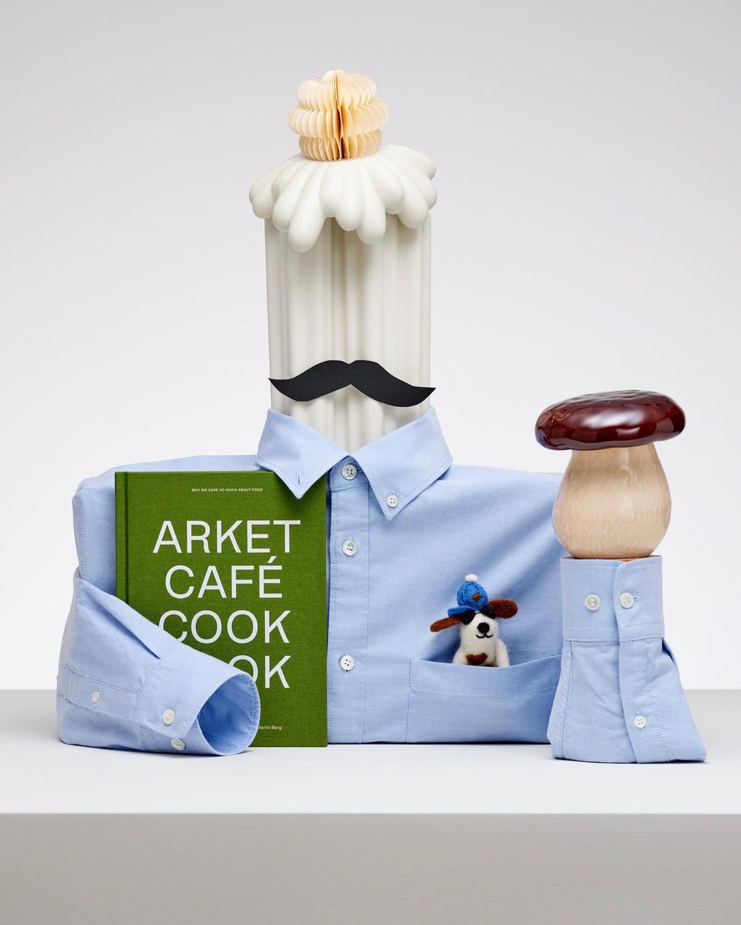 Still life of a white pillar with a black moustache, a blue collared shirt, a cookbook, and a mushroom on a white surface.