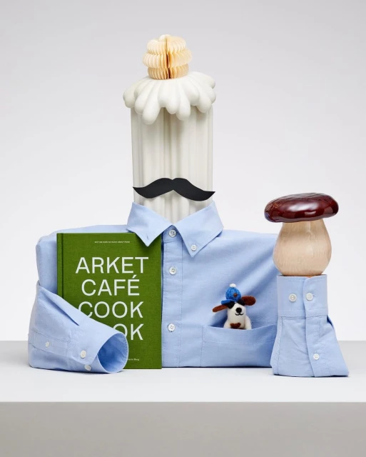 Still life of a white pillar with a black moustache, a blue collared shirt, a cookbook, and a mushroom on a white surface.