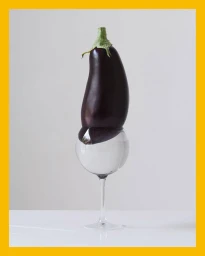 An eggplant balanced vertically on top of a water-filled glass goblet against a white background with a yellow border.