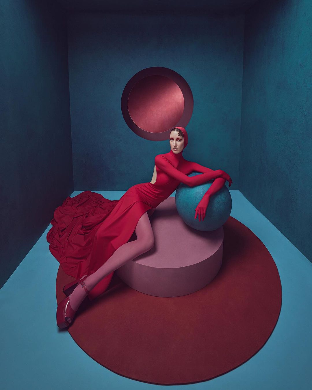 A fashion portrait of a female model in a red dress, posing on a pink platform with a blue sphere, against a blue backdrop with a red circle.