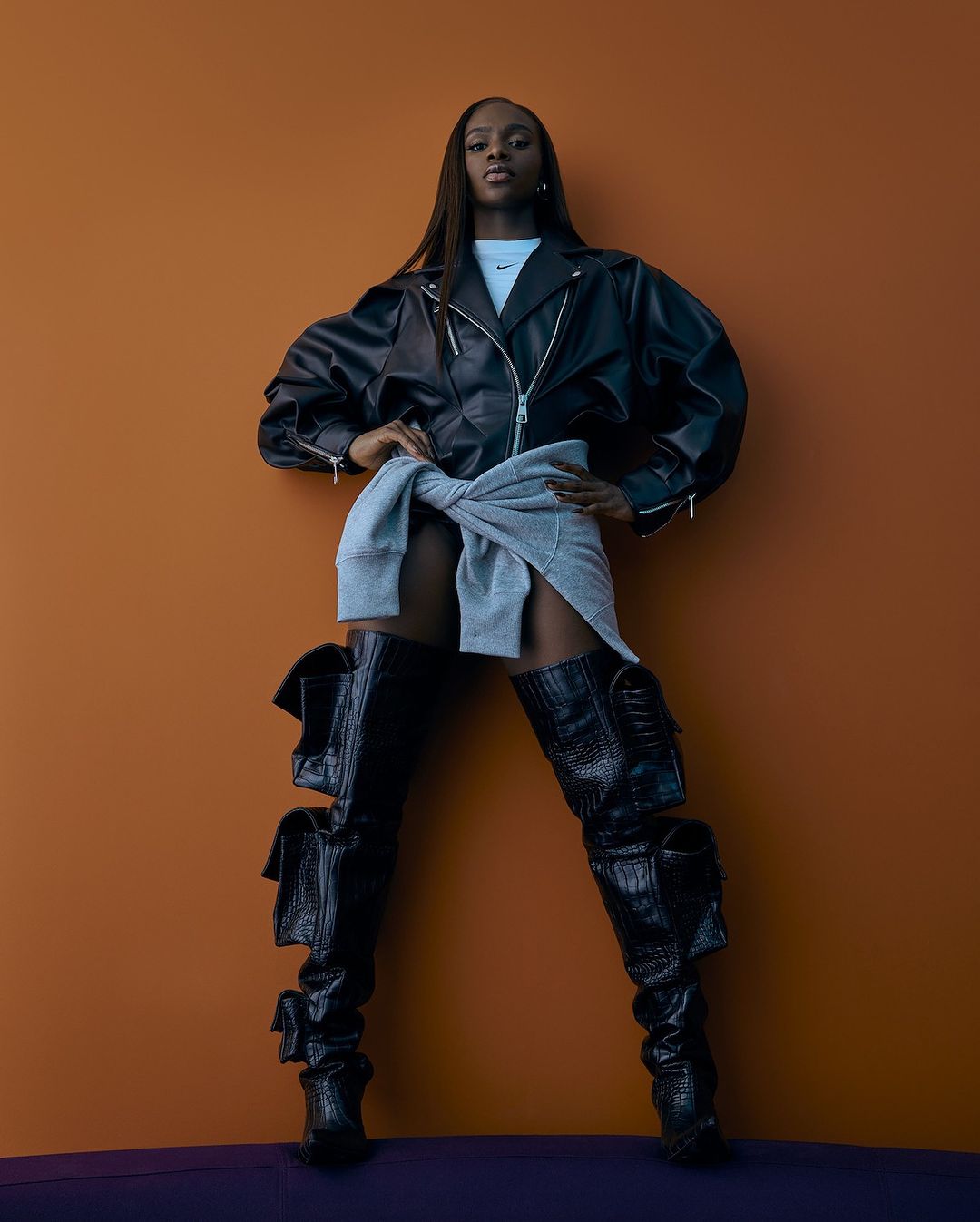 Stylish woman in black leather jacket and thigh-high boots poses against an orange backdrop with a gray sweatshirt tied around her waist.