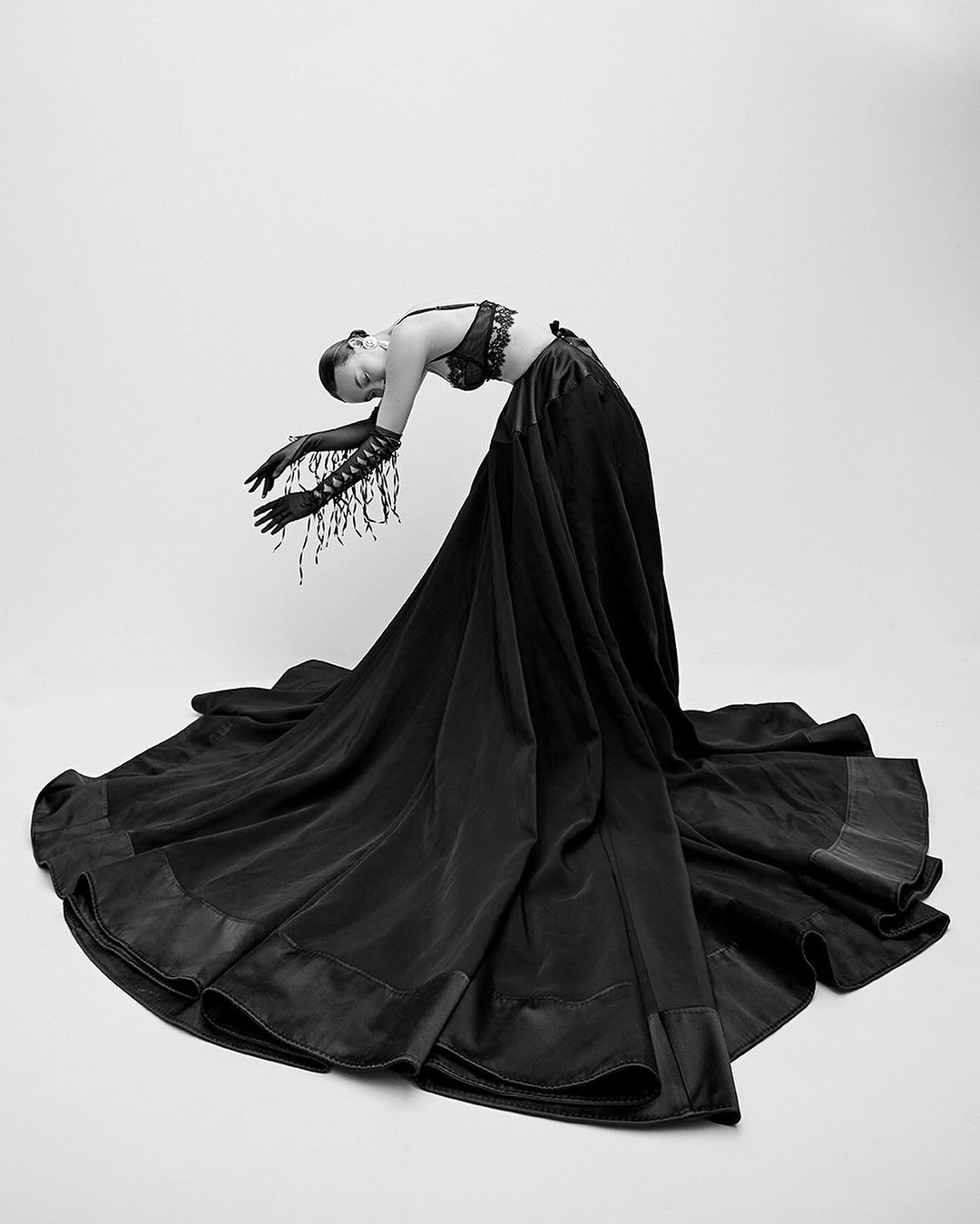 An adult female model wearing a dark two-piece outfit and long fringed gloves is arching backward against a white backdrop in a studio shot.