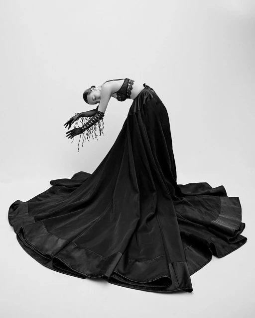 An adult female model wearing a dark two-piece outfit and long fringed gloves is arching backward against a white backdrop in a studio shot.