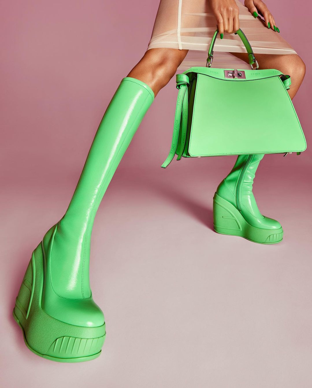 Fashion shot of a woman wearing green knee high boots and carrying a green handbag on a pink background, studio lighting.