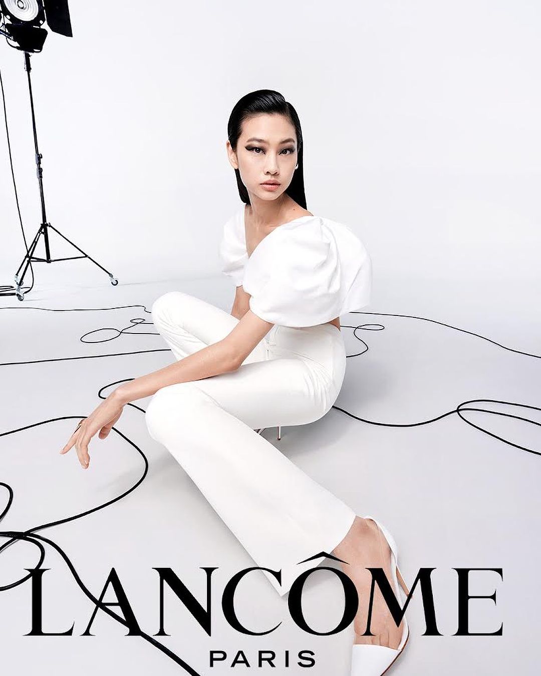 Elegant Asian model in white outfit poses in a photo studio against a white background with visible studio lighting equipment and Lancome logo.