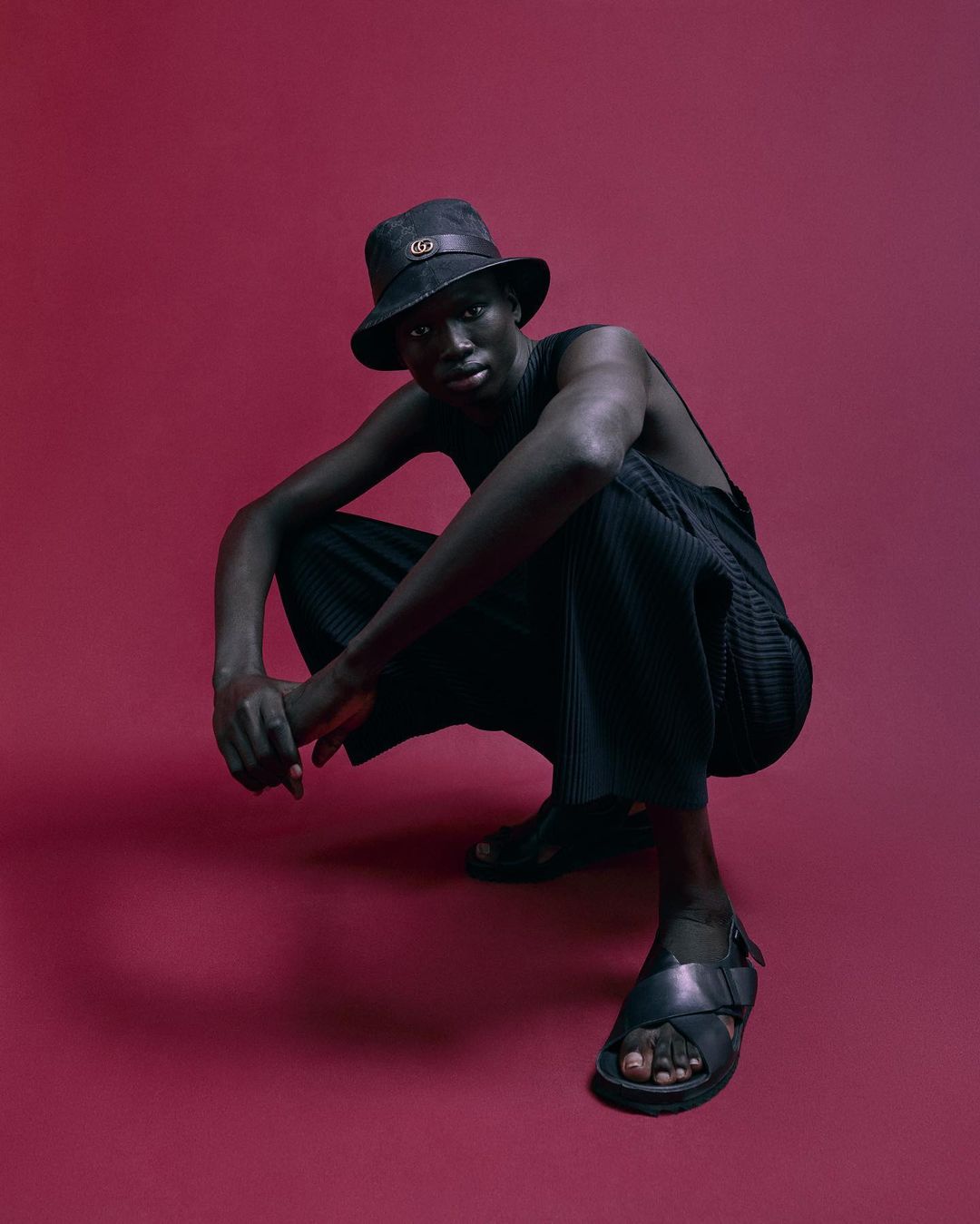 An adult male in a black bucket hat and sandals is crouching against a red background in a studio setting, looking at the camera.