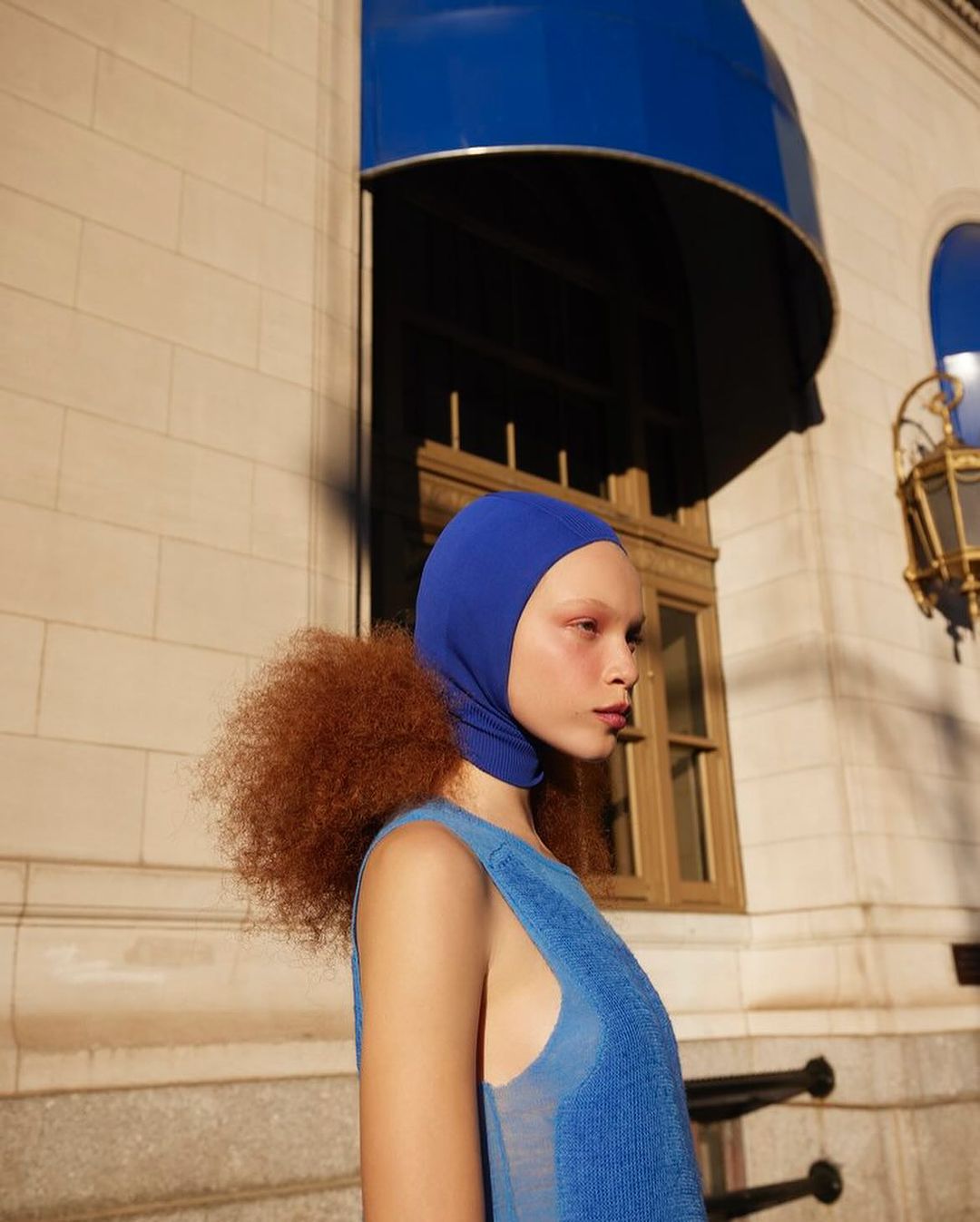Fashionable young woman in blue knitwear and hood poses outdoors in front of a beige building with blue awnings. Modern and striking look.