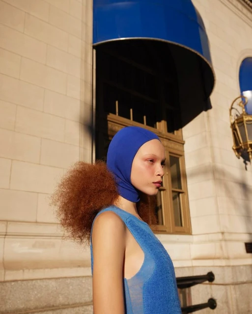 Fashionable young woman in blue knitwear and hood poses outdoors in front of a beige building with blue awnings. Modern and striking look.