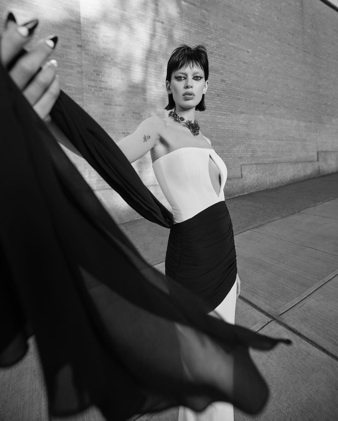 Model in a strapless two-tone black and white dress holding a flowing dark fabric scarf in an urban setting by Carla Guler