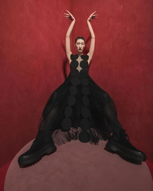 Woman in black dress with circular patterns and black boots posing in front of red wall and pink circular platform.