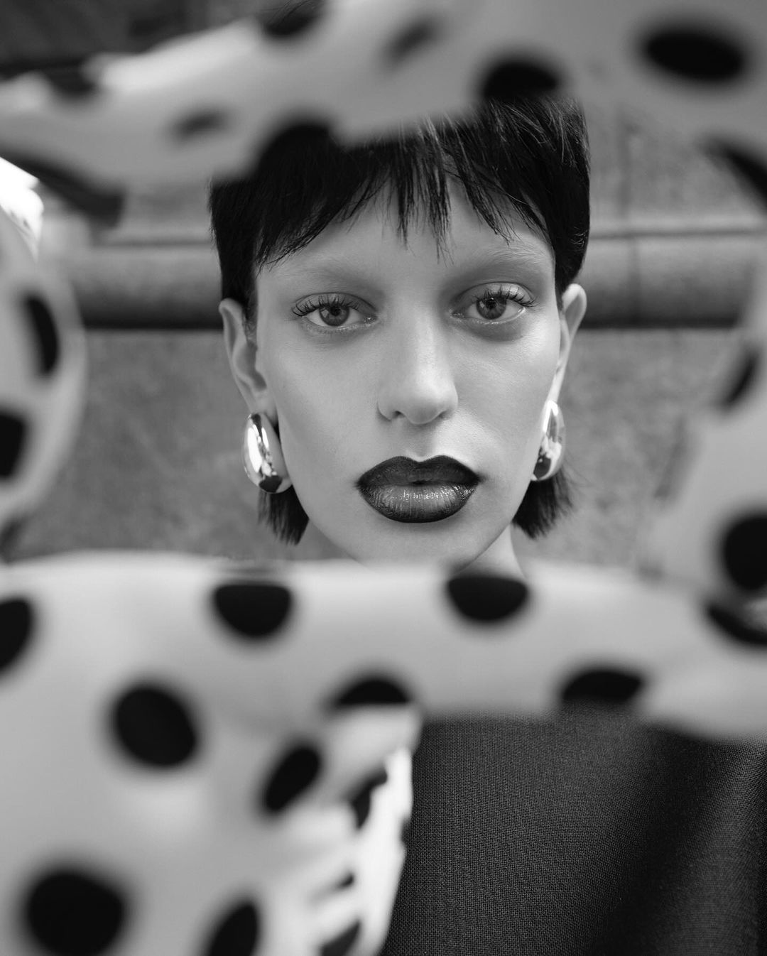 Close-up shot in black and white of a model with dark lipstick and hoop earrings partially obscured by fabric with black polka dots.