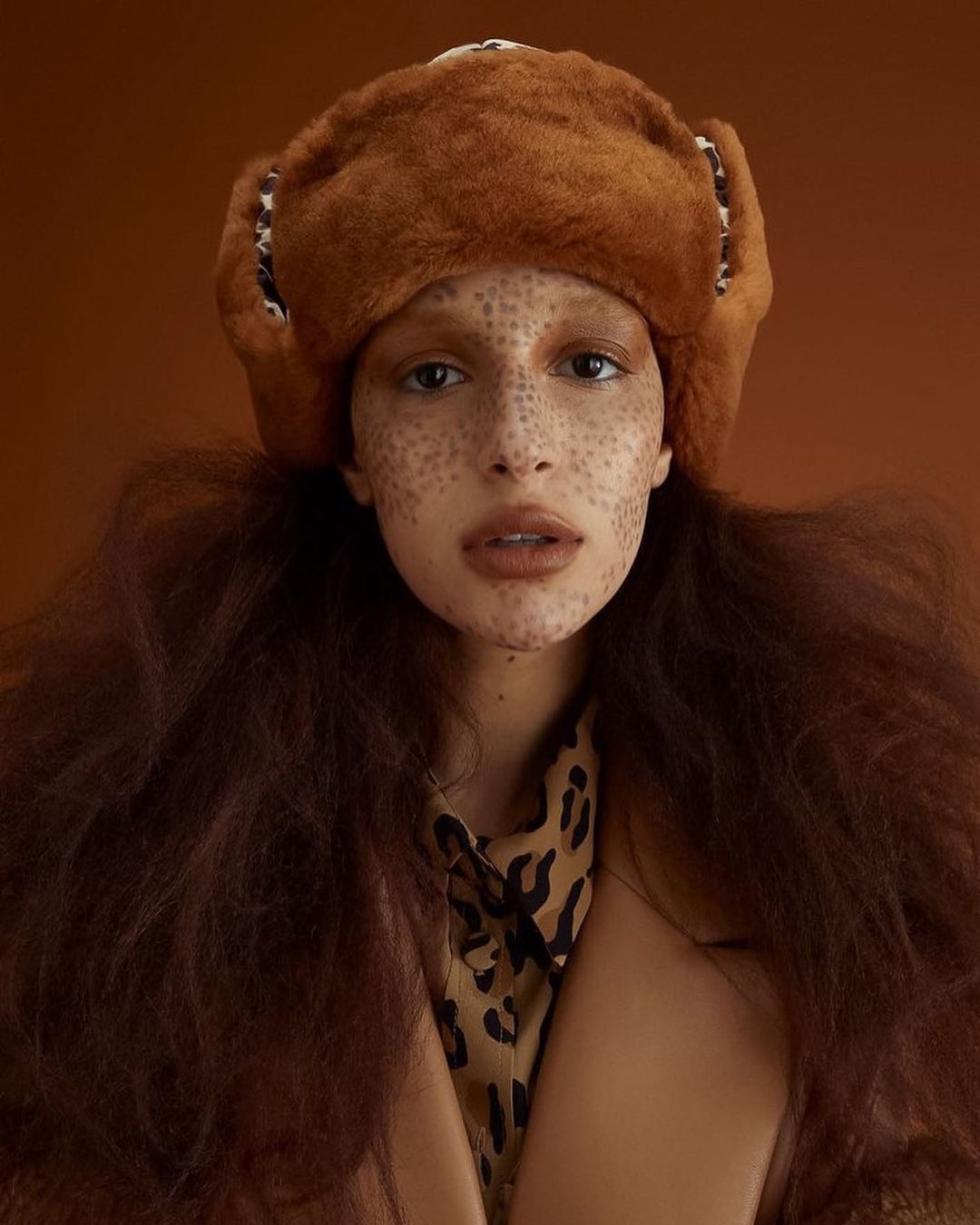 Portrait of a young woman with freckles wearing a fur hat and a patterned shirt against a brown background.
