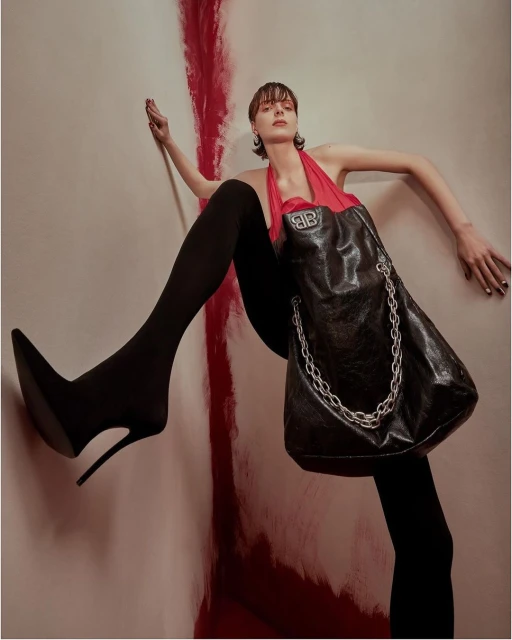Fashion shot: a woman in black tights and heels, posing with a designer bag, against a painted wall with shades of red and beige.