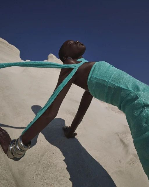 Fashion shot of an African woman in a shimmering teal dress, lying on a white sand dune under a clear blue sky, emphasizing style and pose.