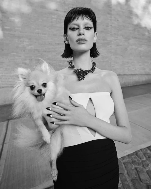 A woman in a strapless top and necklace holds a fluffy dog, with a textured concrete-like wall behind them in a black and white studio shot.