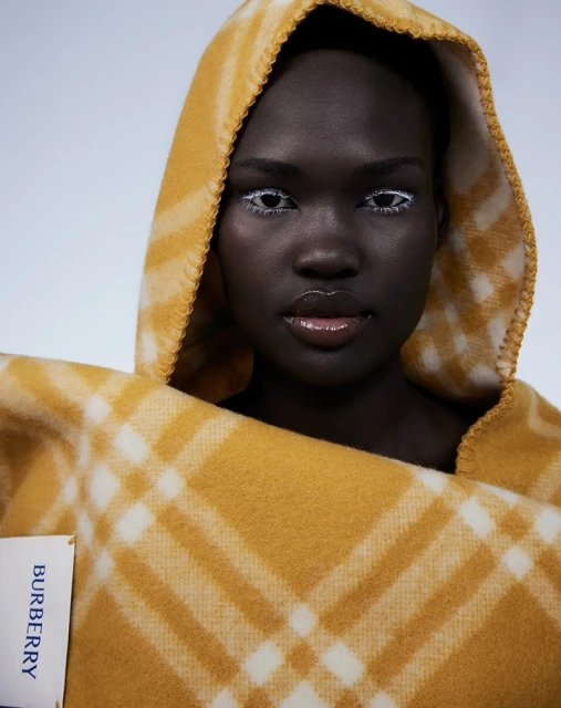 Close-up of a Black female fashion model wearing white eyeliner and a Burberry plaid hood with a matching wrap in a studio setting.