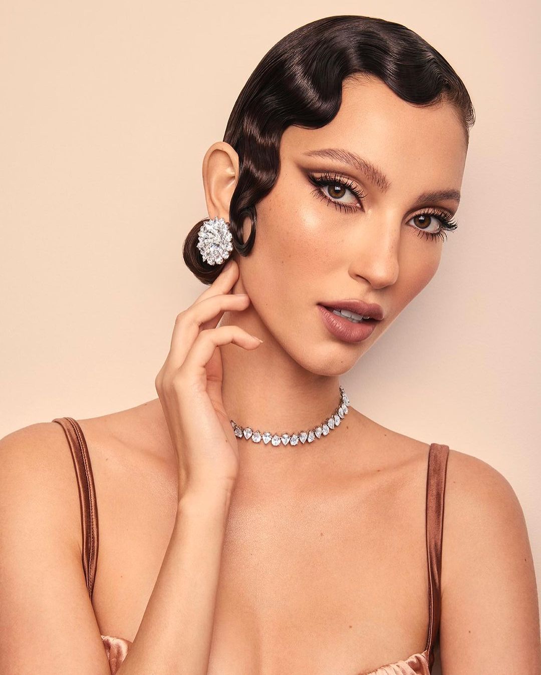Close-up of a woman with dark, waved hair, wearing statement earrings and a necklace, against a pale backdrop. Beauty, jewelry focus.